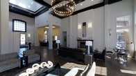 Located at 2234 South Trenton Way in Denver, Colorado, this single-floor space makes a striking first impression with its dramatic, soaring ceilings and bold coffered beam detailing finished in deep espresso tones. A grand circular chandelier serves as a central focal point, casting warm ambient light across the expansive main lounge area.

Floor-to-ceiling arched windows dominate the front-facing wall, flooding the interior with abundant natural light and offering clear views of the surrounding neighborhood. The open layout features multiple distinct seating arrangements, anchored by dark hardwood flooring and layered area rugs that define each zone with purpose and elegance.

A refined fireplace with a classic mantel adds warmth and architectural character to one side of the room, flanked by tasteful wall sconces and decorative accents. The furnishings throughout are contemporary and carefully curated, combining dark leather sofas, sculptural accent pieces, and coordinating tables to create a cohesive, upscale aesthetic.

Beyond the main lounge, an adjacent area — likely used for dining or meetings — is visible through structural columns, hinting at the space's multifunctional design. The overall ambiance is sophisticated and welcoming, blending modern design sensibilities with timeless architectural details to create an impressive and functional gathering environment.