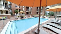 This outdoor courtyard amenity space is designed for relaxation and social gatherings, enclosed by the warm earth-toned stucco facades of a surrounding multi-story building. The centerpiece is a generously sized swimming pool with clear turquoise water, framed by a classic brick-paver deck that adds texture and visual warmth to the environment. Ample lounge chairs are arranged throughout the deck, accented by vibrant orange and yellow market umbrellas that provide shade and a lively pop of color.

Adjacent to the main pool, a bubbling hot tub offers a dedicated space for relaxation, with easy step-in access via stainless steel handrails. Nearby, a built-in outdoor BBQ station equipped with multiple grills serves as a social hub, ideal for outdoor cooking and entertaining. Colorful cushioned seating areas are strategically placed around the courtyard, encouraging conversation and leisure.

The courtyard's enclosed nature creates a semi-private, resort-like atmosphere, sheltered from the outside environment while remaining open to natural light and fresh air. Lush potted greenery softens the hardscape, contributing to the overall inviting and well-appointed ambiance of this communal outdoor space.