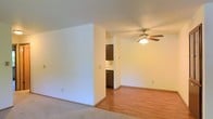 This single-floor residence offers a practical and comfortable layout designed to maximize the use of its open living space. The main living area is finished with soft carpeting, creating a warm and inviting atmosphere, while the adjoining area transitions to smooth hardwood-style flooring that extends toward the kitchen. A ceiling fan with an integrated light fixture is centrally positioned overhead, providing both air circulation and ambient lighting throughout the space.

A partial wall divider adds subtle definition between the living and kitchen areas without fully enclosing either space, preserving an open and airy feel. The kitchen is equipped with dark cabinetry and includes a built-in appliance, likely a dishwasher, tucked neatly beneath the counter. A glimpse of additional upper cabinetry and a wall outlet suggest a functional and well-organized cooking space.

The walls throughout are finished in a neutral, light tone that enhances the brightness of the interior and contributes to a clean, understated aesthetic. The overall layout is straightforward and efficient, making it well-suited for everyday living. The seamless flow between rooms creates a cohesive environment that feels open and easy to navigate.