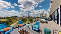 Located at 947 Myers Street in Richmond, VA, this expansive rooftop terrace offers a dynamic outdoor amenity space with sweeping panoramic views of the Richmond skyline and surrounding landscape. The deck features a glass railing system along its perimeter, ensuring unobstructed sightlines across the city while maintaining an open, airy feel throughout.

The terrace is thoughtfully organized into several distinct zones. A partially covered lounge area features an outdoor fireplace with a wall-mounted television, comfortable gray seating, and a ceiling fan, creating a comfortable retreat in varying weather. Nearby, a fire pit surrounded by colorful blue Adirondack chairs serves as a central gathering point, ideal for relaxed socializing.

An outdoor kitchen equipped with multiple grills provides ample space for cooking and entertaining. Adjacent to it, a dining area with bright yellow chairs and white tables is set against a striking accent wall of natural wood and vertical greenery panels, adding a fresh, organic element to the space.

String lights are strung across the length of the terrace, lending a warm, festive ambiance during evening hours. Additional dining and seating arrangements are positioned along the glass railing to maximize enjoyment of the views. The light-toned wood decking ties the entire space together, creating a cohesive and inviting rooftop destination.
