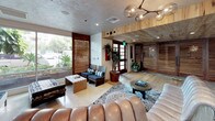 This single-floor space presents a thoughtfully curated interior that merges artistic sensibility with casual, lived-in comfort. The design language is defined by exposed rustic wood ceiling beams contrasting against white shiplap ceilings and polished concrete flooring, creating a warm yet contemporary aesthetic that carries consistently throughout the space.

The layout unfolds across several interconnected zones. A gallery-like area showcases large-scale artwork on crisp white walls, including a bold red portrait piece, framed by steel-framed glass doors that lend an industrial touch. Natural light filters in through full-height windows and slatted wooden shutters, contributing to the bright, airy character of the interiors.

A recreation area features a foosball table, a suspended rope-hung boat-style daybed, a surfboard display, and vibrant orange canvas art, reinforcing an eclectic, resort-inspired atmosphere. Nearby, a more intimate lounge nook offers a compact sofa with yellow accent pillows, a backgammon table, a marble-topped coffee table, and a decorative wall hanging, creating a relaxed retreat.

The main lounge area is anchored by plush leather seating, a sculptural branch chandelier, warm wood paneling, and generous greenery, bringing together the space's natural and artistic elements cohesively.
