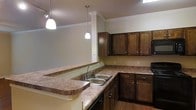 This single-floor residence offers a thoughtfully designed layout that combines functionality with classic style. The kitchen serves as a standout feature, showcasing rich dark wood cabinetry with brushed nickel hardware that provides generous storage throughout. The countertops display an earthy, marbled laminate surface that extends across an L-shaped configuration, offering ample workspace for cooking and meal preparation. A double-basin stainless steel sink is centrally positioned along the counter, adding a practical and polished touch. The kitchen opens to an adjacent area through a peninsula-style breakfast bar, enhancing the sense of connectivity and flow between spaces. Two elegant pendant lights hang above, casting warm, focused illumination that complements the overall ambiance. Crown molding trims the ceiling throughout, lending a refined architectural detail to the space. A decorative column with crown molding detail further accentuates the transition between the kitchen and the adjoining living area. Hardwood-style flooring grounds the space with warmth and durability. The neutral wall tones provide a versatile backdrop that pairs well with the rich cabinetry tones, creating a cohesive and inviting interior environment throughout this well-crafted residence.
