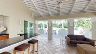 This single-level covered outdoor pavilion is designed for comfortable tropical living and entertaining. The structure features an elevated, white-painted wood-beam ceiling with exposed rafters, lending the space a clean, classic coastal character. Ceiling fans are mounted overhead to ensure airflow and comfort throughout the area.

The flooring is composed of large-format travertine tiles in a neutral stone finish, adding an elegant yet durable surface suited to outdoor use. A built-in bar counter with a dark granite top runs along one side of the pavilion and is accompanied by woven rattan bar stools with padded leather seats, creating a casual gathering spot. A seating area with a leather armchair and a small side table provides a relaxed lounge area nearby.

Structural columns define the open perimeter of the pavilion, framing unobstructed views of the surrounding tropical landscape. Lush palm trees, manicured planters, and a pool area are fully visible from within the space, seamlessly connecting the interior to the outdoors. Large sliding glass doors on one side suggest access to an adjoining interior space. The overall ambiance is bright, airy, and resort-like, making this pavilion a standout feature well-suited for both daytime relaxation and evening gatherings.