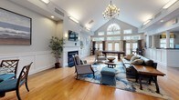 Located at 184 Federal Way in Johnston, Rhode Island, this single-floor community clubhouse offers a thoughtfully designed amenity space that balances elegance with functionality. The centerpiece is a grand common room boasting soaring vaulted ceilings, warm hardwood floors, and an arched window array that floods the space with natural light. A classic chandelier hangs above multiple seating arrangements, while a fireplace and wall-mounted television anchor one side of the room. Wainscoting along the walls and decorative columns add refined architectural detail throughout.

Adjacent to the common room is a formal dining nook with a glass-top table, chair rail molding, and coastal-themed artwork. A well-equipped kitchenette nearby features dark granite countertops, subway tile backsplash, a stainless steel dishwasher, and a commercial-grade coffee station.

The fitness center is divided into two well-lit areas — one dedicated to cardio equipment including treadmills, ellipticals, and stationary bikes, and another featuring a multi-station strength machine and free-standing equipment, both accented by vibrant abstract murals.

A carpeted lounge and business area rounds out the space, offering teal accent chairs, contemporary artwork, and a built-in counter workspace — providing residents a relaxed yet productive environment.