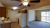 This single-floor residence features an open-concept design that connects the kitchen and living area, creating a functional and cohesive layout. The kitchen is equipped with warm, medium-toned wood cabinetry mounted both above and below the counter level, providing ample storage. White appliances, including a full-size refrigerator and an electric range, complement the space, which features durable tile flooring in a classic checkered pattern. A ceiling-mounted light fixture provides additional task lighting in the kitchen area.

A decorative ceiling fan with a built-in light fixture is centrally positioned overhead, serving both the kitchen and adjacent living area. The transition from the kitchen's tile flooring to the carpeted living space is clearly defined, offering a comfortable and distinct zone for relaxation. The living area features neutral walls, standard baseboards, and an open entryway that enhances the sense of flow throughout the unit. The textured ceiling and HVAC vent indicate standard climate control infrastructure. Overall, the space maintains a straightforward, practical design with a warm, neutral color palette throughout.