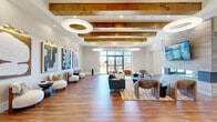 Located at 2901 Elmwood Dr in Harrisburg, PA, this thoughtfully designed single-story community facility blends contemporary aesthetics with warm, organic materials to create an inviting and functional gathering space. The expansive living room serves as the centerpiece, featuring rich hardwood flooring, exposed wooden ceiling beams, and dramatic circular pendant lights that draw the eye upward. Large floor-to-ceiling windows flood the space with natural light and offer open views of the surrounding landscape, while a sleek stone-clad fireplace with a mounted flat-screen TV anchors the room. Curated modern artwork lines the walls, complementing the carefully selected lounge furniture arranged throughout the space. The hallways echo the design language of the main area, with wooden beam accents and gallery-style art displays creating a cohesive flow between rooms. The well-equipped exercise room is bathed in natural light from expansive black-framed windows and features durable rubber flooring, a vaulted ceiling with pendant lighting, and a full complement of cardio and strength training equipment, including treadmills, ellipticals, a rowing machine, and a cable machine. A cozy media room with dark, moody walls, recessed lighting, and a large wall-mounted screen provides an intimate setting for entertainment. The dining area rounds out this comprehensive amenity space, offering residents a versatile environment for socializing and daily activities.
