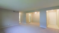 This single-floor space presents a straightforward, functional layout with a notably open feel. The main living area is finished with wall-to-wall carpet in a neutral tone, contributing to a clean and understated aesthetic throughout. Soft grey-toned walls provide a calm, neutral backdrop that works well with a variety of furnishing styles.

A wide open passthrough or recessed alcove connects the living area to an adjacent room, which is equipped with a ceiling fan and light fixture, offering both air circulation and overhead illumination. This secondary space is versatile and could serve as a dining area or additional living zone. A full-size refrigerator is visible through a further opening, indicating a connected kitchen area just beyond.

To the left, a separate passageway leads toward what appears to be an additional room or hallway, suggesting a multi-room configuration despite the single-floor footprint. HVAC vents are visible in the ceiling, ensuring climate control throughout. White baseboards trim the perimeter of each room, adding a crisp contrast to the carpeted floors. Overall, the space offers a practical, neutral canvas well-suited for customization.