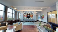 This single-floor amenity lounge is thoughtfully designed to blend sophistication with functionality. The open-plan layout is anchored by rich hardwood flooring that contrasts elegantly with light gray walls, creating a warm yet modern aesthetic. A centerpiece billiard table in a sleek gray finish sits atop a large area rug, serving as the focal point of the recreational zone. Overhead, a pair of starburst-style chandeliers cast ambient light, complementing the recessed lighting integrated into the tray ceiling above.

At the rear of the space, a built-in bar and entertainment area features a mounted flat-screen television flanked by vibrant, colorful wall panels that add a dynamic visual accent. White contemporary seating surrounds the bar, creating an inviting social hub. Large floor-to-ceiling windows along the perimeter flood the space with natural light and provide views of the surrounding greenery.

Bold abstract artwork adorns the walls throughout, reinforcing the space's contemporary character. Additional seating, including a cream upholstered armchair with nailhead trim, contributes to the lounge's relaxed yet polished ambiance. A decorative floral arrangement adds a refined finishing touch.