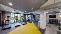 Located at 401 S Justison St in Wilmington, Delaware, this fitness center offers a thoughtfully designed exercise space across a single floor. The room features a modern, contemporary aesthetic with a combination of gray rubber flooring and vibrant yellow matting that delineates the open stretching and functional training zone from the equipment areas.

The cardio section is well-appointed with multiple treadmills, elliptical trainers, and stationary bikes, each equipped with individual monitors for an engaging workout experience. Large floor-to-ceiling windows line one wall, flooding the space with natural light and providing views of the surrounding greenery — an energizing backdrop for any workout.

The strength training area includes a selection of cable machines, a weight bench, and additional selectorized equipment, all arranged to allow comfortable circulation throughout the space. Full-length mirrors along one wall enhance the sense of openness while allowing users to monitor their form. Warm wood-paneled accent walls add a refined, modern touch to the interior design.

Overhead, ceiling fans and recessed lighting ensure the space remains well-ventilated and evenly lit. A beverage refrigerator and waste receptacles are conveniently placed for user comfort. Glass-paneled entry doors connect the fitness center to the adjacent building, making it easily accessible. The overall ambiance is clean, energetic, and conducive to focused exercise.