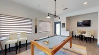 This single-floor amenity space is thoughtfully designed to serve as both a recreational and functional hub. The centerpiece of the main game room is a handsome wood-framed billiard table with a slate-gray felt surface, positioned beneath a striking multi-pendant industrial-style light fixture that provides focused illumination. A wall-mounted cue rack keeps equipment neatly organized nearby. The room is brightened by multiple windows dressed with layered zebra-style blinds that allow for flexible light control, while the floors feature durable wood-look tile throughout. A colorful abstract area rug anchors the billiard table, adding a lively design element to the space. High-top bar seating and a small round bistro table offer comfortable spots for spectators or casual socializing. Vibrant pop-art style wall prints lend the space a playful, modern personality. A secondary alcove area provides a compact counter with a small appliance setup, ideal for light refreshments. Flowing from the game room is a dedicated business center corridor, lined with a long built-in desk, computer workstations, and rolling task chairs — offering a quiet, productive environment. The overall ambiance is clean, contemporary, and welcoming, with bright white walls, teal accent baseboards, and well-coordinated furnishings throughout.