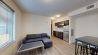 This single-floor unit offers a clean, modern layout designed for comfortable everyday living. The open-concept living area is anchored by light hardwood-style flooring that extends throughout the space, creating a cohesive and bright atmosphere. A large window fitted with horizontal blinds allows natural light to fill the living room, which comfortably accommodates a sectional sofa and a coffee table.

Adjacent to the living area, the kitchen is efficiently laid out and features dark espresso-toned cabinetry — both upper and lower — paired with a white countertop and a stainless steel appliance suite, including a refrigerator, over-the-range microwave, dishwasher, and a cooktop with a single-basin sink.

The unit includes two bedrooms, each finished with the same light flooring and neutral walls. One bedroom features a desk positioned beneath a window, providing natural light for a workspace. The other bedroom includes a dresser and a tall storage unit, with easy access to the hallway. Both rooms offer a functional and comfortable layout.

The full bathroom is finished in white tile flooring and includes a tub-shower combination, a single-sink vanity with ample storage, and a large mirror, maintaining the unit's clean and practical aesthetic throughout.