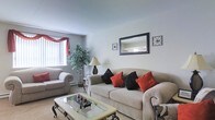 This single-floor space offers a warm and inviting atmosphere centered around a thoughtfully arranged living room. The living area is furnished with a plush, neutral-toned sofa and matching armchair, accented with bold red and dark throw pillows that add a striking pop of color to the room. A glass-top coffee table with ornate metal detailing serves as a stylish focal point, complementing the overall classic décor. Natural light filters in through a large window dressed with rich red curtains and vertical blinds, brightening the space and creating an airy feel. The walls are kept in a clean, neutral tone, providing a versatile backdrop for the decorative elements throughout. A framed wall mirror flanked by metal wall art adds depth and dimension to the room, while a tall indoor plant introduces a natural, refreshing touch. The flooring features light-toned carpeting that adds warmth and comfort underfoot. A baseboard heating unit along the wall ensures year-round comfort. The overall design strikes a balance between functionality and classic elegance, making the living area a welcoming retreat suited for both relaxing and entertaining.