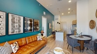 This thoughtfully designed single-floor residence showcases a contemporary open-concept layout that seamlessly connects the living, dining, and kitchen areas. The living room immediately draws the eye with its striking teal accent wall, light wood flooring, and floor-to-ceiling glass doors that invite abundant natural light and provide access to a private balcony. The kitchen is a standout feature, outfitted with warm wood cabinetry, dark granite countertops, a herringbone-patterned tile backsplash, and a full suite of stainless steel appliances including a side-by-side refrigerator, range, and dishwasher. A wide waterfall-edge island with bar seating bridges the kitchen and living areas, making the space ideal for both cooking and entertaining. The dining area offers a cozy round table setting complemented by an eclectic wall display of woven basket art. The bedroom is a calm retreat, featuring large windows with ample natural light, soft neutral tones, and generous wall space for artwork. The bathroom continues the cohesive design with light wood vanity cabinetry, granite countertops, and a spacious glass-enclosed tile shower. Light wood flooring flows consistently throughout, tying the entire residence together with warmth and style.