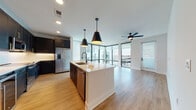 Located in the Midtown Tampa district, this single-floor residence offers a thoughtfully designed open-concept layout filled with contemporary finishes throughout. The kitchen serves as the heart of the home, showcasing dark navy shaker-style cabinetry with gold and matte black hardware, a large center island with a white quartz countertop, an undermount sink with a matte black faucet, and a full suite of stainless steel appliances including a built-in microwave, electric range, and French door refrigerator. Stylish pendant lighting hangs above the island, complementing the under-cabinet lighting and recessed ceiling fixtures. The living area extends seamlessly from the kitchen, highlighted by floor-to-ceiling glass windows and a sliding door that opens to an outdoor balcony, flooding the space with natural light. Light-toned wood-look flooring runs consistently throughout the unit, tying each room together. The bedrooms are bright and airy, each featuring ceiling fans, large windows with views of the surrounding neighborhood, and ample closet space, including a well-organized walk-in closet. The bathrooms are finished with dark navy vanities, white quartz countertops, LED-backlit mirrors, and matte black fixtures, with one bathroom offering a tub-shower combination and the other a spacious vanity with a tall linen tower.