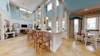 Located at 501 Burke Dr in Hinesville, Georgia, this single-story community clubhouse is a beautifully appointed gathering space designed for both comfort and socializing. The interior is defined by dramatically soaring ceilings accented with soft teal-blue walls above a neutral wainscoted lower half, creating an airy and refined atmosphere throughout.

The main lounge area features two generously sized sofas arranged around a round cane-accented coffee table, all set atop a bold patterned area rug. A wall-mounted TV and decorative sunburst mirror complete the inviting sitting room. Overhead, an elegant chandelier adds a warm focal point to the space.

A central bar-height island with granite countertops and white beadboard paneling serves as a stylish divider between the lounge and kitchen areas. The full kitchen is equipped with white cabinetry, dark wood countertops, a stainless steel sink, and ample prep space, with views opening directly into the social areas.

The open gathering hall features travertine tile flooring, tall clerestory windows that flood the space with natural light, and multiple seating arrangements with upholstered armchairs and side tables. A separate dining nook with round tables and cross-back chairs offers views of the outdoor pool area through large draped windows with direct exterior access.