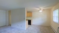 This single-floor space features an open-concept design that connects the living area to the kitchen, creating a functional and cohesive layout. The living area is finished with wall-to-wall carpet in a neutral tone, providing a comfortable underfoot feel, while a ceiling fan with an integrated light fixture serves as both a practical and central overhead feature.

A half-wall partition subtly defines the transition between the living space and the kitchen without fully closing off the two areas, maintaining an airy, connected feel. The kitchen features light wood cabinetry offering ample storage, a countertop workspace, a built-in sink, and a black dishwasher positioned beneath the counter. The kitchen floor transitions to a hard surface, distinguishing it from the carpeted living area.

The walls throughout are painted in a clean, light gray-white tone, contributing to a bright and neutral ambiance. The overall design is straightforward and practical, with consistent finishes that offer a blank canvas for personalization. The space is well-suited for everyday living with its efficient and accessible layout.