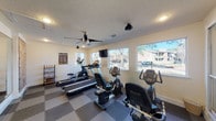 This fitness center is a dedicated, single-floor workout facility designed to support a well-rounded cardio routine. The space is outfitted with a variety of cardio equipment, including treadmills, upright and recumbent stationary bikes, and an elliptical trainer, all positioned to allow comfortable use throughout the room. Interlocking foam floor tiles in a gray and tan checkered pattern cover the entire floor, providing both cushioning and durability underfoot. A ceiling fan with a sleek black finish is centrally mounted, ensuring consistent air circulation throughout workouts. A wall-mounted flat-screen television is positioned for easy viewing from multiple machines. Large windows line one wall, inviting an abundance of natural light into the space while offering views of the surrounding outdoor area. A full-length mirror on one wall enhances the sense of openness and allows users to monitor their form. A small shelving unit near the entrance holds amenity items, and a posted fitness center rules board keeps guidelines visible. Recessed lighting and a textured ceiling contribute to the room's clean, functional aesthetic. The facility is accessed through a distinctive red-framed glass door with decorative sidelights, adding a touch of character to the entrance.