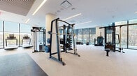 This contemporary fitness center is thoughtfully designed to support a wide range of exercise disciplines within a single, well-organized level. The space is divided into distinct functional zones, each finished with clean, modern detailing and bathed in abundant natural light from expansive floor-to-ceiling windows that frame views of surrounding greenery and the urban streetscape beyond.

The primary strength training area is generously proportioned, accommodating multiple cable machines, power racks, a Smith machine, and a comprehensive dumbbell station with built-in shelving for organized storage. Neutral-toned carpet flooring anchors this zone, complementing the white ceilings and recessed linear lighting overhead. Structural columns are integrated seamlessly into the layout, contributing to the space's refined aesthetic without interrupting the flow of equipment.

A dedicated cardio zone lines the perimeter near the floor-to-ceiling glazing, housing treadmills, an elliptical, a rowing machine, and a stationary cycling bike. This area benefits particularly from the panoramic views and natural daylight.

A separate studio room offers hardwood flooring, full-length mirrors along one wall, mounted flat-screen displays, and an open layout suitable for yoga, cycling, or group fitness classes. The overall ambiance throughout is bright, clean, and professionally appointed.