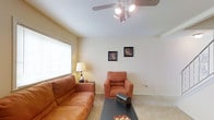 This two-floor residence offers a welcoming and functional layout designed for everyday comfort. The living area sets a relaxed tone with its warm neutral color palette, featuring soft beige walls and plush wall-to-wall carpeting that adds a cozy feel underfoot. A ceiling fan with integrated lighting serves as a practical central fixture, ensuring good air circulation and illumination throughout the room. A large window fitted with horizontal blinds allows natural light to filter into the space while maintaining privacy, contributing to the bright and airy atmosphere. The living room is well-proportioned, providing ample space for seating and relaxation. Clean, smooth walls offer a versatile backdrop suitable for personalization. The overall design of the main living area emphasizes simplicity and ease of living, with a layout that flows naturally. Spanning two floors, the home provides clear separation between living and private spaces, making it a practical and comfortable residence. The understated finishes and neutral tones throughout create a calm, inviting ambiance that is both timeless and adaptable to a variety of interior styles.