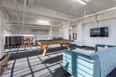 Located at 1028 Second Ave S in Birmingham, Alabama, this single-floor amenity space showcases a compelling blend of industrial character and modern comfort. The open layout is defined by exposed white-painted structural beams, visible ductwork, and high ceilings that lend an airy, warehouse-inspired aesthetic throughout.

Natural light pours generously through a row of large, multi-pane industrial windows along one wall, casting dramatic sunlight patterns across the mixed flooring — a combination of patterned carpet zones and raw, patinated concrete that highlights the space's original character.

The main lounge area features a teal mid-century-style sofa paired with accent seating, centered around a wall-mounted flat-screen television. A full-size pool table with a green felt surface serves as a focal point of the recreation zone, complemented by a ping pong table nearby, making the space well-suited for social gatherings and entertainment.

A raised mezzanine-style section, bordered by sleek black metal railings, offers a more intimate seating area with a bar-height table and chairs — ideal for small group conversations or casual meetings. Additional café-style seating is available in a separate nook framed by exposed structural columns. A teal accent door adds a pop of color, contributing to the space's vibrant, welcoming atmosphere.