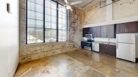 Located at 144 E Fourth St in Burlington, NC, this single-floor loft showcases an industrial-chic aesthetic with soaring ceilings, exposed brick walls, and polished concrete floors throughout. The open living and kitchen area is anchored by floor-to-ceiling windows that invite abundant natural light and outdoor views, complemented by a ceiling fan and track lighting overhead. The kitchen features stainless steel appliances, dark cabinetry, and granite countertops. The bedroom offers clerestory windows and double-door closet storage. The full bathroom includes a tub/shower combo, granite vanity, and dark wood cabinetry. A hallway connects the spaces cohesively, completing this urban, loft-style residence.