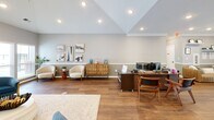 Located at 4900 Chandler Ridge Circle in Raleigh, NC, this single-story space offers a well-organized layout across a generously sized floor plan. The living and kitchen area serves as the heart of the space, highlighted by a striking full-height brick fireplace flanked by large windows that flood the room with natural light. The vaulted ceiling adds a sense of openness, while warm hardwood flooring flows consistently throughout. The kitchen features warm-toned wood cabinetry, white countertops, stainless steel appliances including a French door refrigerator and dishwasher, and a peninsula that defines the transition between kitchen and living areas. The hallway entry provides a bright, welcoming reception area with a ceiling fan, multiple windows, seating, and direct exterior access. An adjacent workspace area features recessed lighting beneath a vaulted ceiling and provides ample room for workstations and seating. The dedicated exercise room is equipped with fitness equipment, motivational wall décor, crown molding, a ceiling fan, and its own exterior door. Together, these rooms create a functional, well-lit, and thoughtfully arranged space suited for a variety of uses.