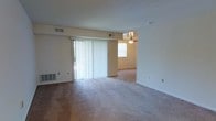 This single-floor space features an open layout connecting two living areas, both finished with wall-to-wall carpeting in neutral tones. The main living area is anchored by a large sliding glass door dressed with vertical blinds, allowing generous natural light to fill the room. A wall-mounted heating unit sits nearby, adding a practical element to the space.

An open passage leads to a secondary room, which is equipped with a ceiling fan with a light fixture and an additional window that further brightens the interior. The continuous carpet flooring and consistent neutral wall color unify both areas, creating a cohesive and understated aesthetic throughout.

Overhead, HVAC vents are integrated into the ceiling, ensuring climate control across the living spaces. The layout is straightforward and functional, with multiple electrical outlets positioned along the walls for everyday convenience. The overall ambiance is light and airy, with the combination of sliding glass door access and multiple windows contributing to a well-lit, open feel throughout the single-floor layout.