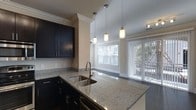 This single-floor residence offers a thoughtfully designed open-concept layout that seamlessly connects the kitchen and living areas. The kitchen is a standout feature, showcasing rich, dark cabinetry that contrasts elegantly with light granite countertops. A double stainless steel sink is positioned at the peninsula island, which also serves as a natural divider between the kitchen and the adjacent living and dining space. Stylish pendant lights hang above the peninsula, providing both task lighting and a decorative focal point. Crown molding runs along the ceilings throughout, adding a refined architectural detail that elevates the overall aesthetic. The living area is bathed in natural light, thanks to a large window and a sliding glass door fitted with vertical blinds that lead to an outdoor area, offering views of lush greenery. The warm, neutral wall tones complement the dark cabinetry and light countertops, creating a cohesive and inviting ambiance. Track lighting provides additional illumination, ensuring the space is well-lit at all times. The combination of upscale finishes and practical design elements makes this residence both functional and visually appealing.