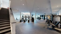 Located at 1340 S Hill St in the heart of Los Angeles, this fitness center offers a generously sized, single-floor exercise space designed to accommodate a full range of workout needs. The facility is laid out across an open, airy floor plan with durable rubber flooring throughout, providing both comfort and functionality for users.

The cardio zone is well-equipped with multiple treadmills, elliptical machines, stationary bikes, stair climbers, and a rowing machine, several of which feature individual display screens for interactive training. Large windows and glass doors allow natural light to pour in, creating an energizing atmosphere, while soaring clerestory windows in the cardio section further amplify the bright, open feel.

The strength training area is thoughtfully arranged with a full rack of dumbbells, multiple cable and resistance machines, and dedicated weight benches, offering options for both isolation and compound exercises. Structural columns delineate zones without disrupting the spacious flow of the room.

A corridor along the staircase leads to an additional equipment area, adding versatility to the overall layout. White walls, recessed lighting, and a clean modern aesthetic give the space a polished, professional appearance. The facility is well-ventilated with visible HVAC systems throughout, ensuring a comfortable environment for all levels of fitness activity.