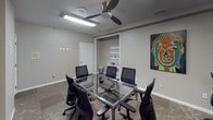 This single-floor office space is thoughtfully designed to support both focused individual work and collaborative meetings. The layout includes a private workstation area furnished with a built-in desk, computer setup, and ergonomic seating, making it well-suited for heads-down productivity. A striking large-scale blue and gold abstract painting anchors the wall, adding a creative touch to the work environment.

The conference room serves as the centerpiece of the space, featuring a sleek glass-top table with a chrome base surrounded by ergonomic mesh chairs, comfortably accommodating multiple attendees. A ceiling fan with integrated lighting keeps the room comfortable during extended meetings. A built-in nook with open shelving provides convenient storage for office supplies and materials, while wall-mounted display rails offer space for notes or visual presentations.

Throughout the office, patterned carpet flooring adds warmth and texture, complementing the neutral gray walls. Colorful artwork, including a vibrant pop-art style portrait and a bold abstract canvas, is displayed across the space, creating an engaging and inspiring atmosphere. The overall ambiance is professional yet creatively stimulating, balancing functionality with distinctive aesthetic character.