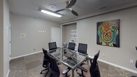 This single-floor office space is thoughtfully designed to support both focused individual work and collaborative meetings. The layout includes a private workstation area furnished with a built-in desk, computer setup, and ergonomic seating, making it well-suited for heads-down productivity. A striking large-scale blue and gold abstract painting anchors the wall, adding a creative touch to the work environment.

The conference room serves as the centerpiece of the space, featuring a sleek glass-top table with a chrome base surrounded by ergonomic mesh chairs, comfortably accommodating multiple attendees. A ceiling fan with integrated lighting keeps the room comfortable during extended meetings. A built-in nook with open shelving provides convenient storage for office supplies and materials, while wall-mounted display rails offer space for notes or visual presentations.

Throughout the office, patterned carpet flooring adds warmth and texture, complementing the neutral gray walls. Colorful artwork, including a vibrant pop-art style portrait and a bold abstract canvas, is displayed across the space, creating an engaging and inspiring atmosphere. The overall ambiance is professional yet creatively stimulating, balancing functionality with distinctive aesthetic character.