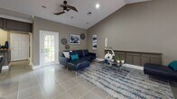 This single-floor space is defined by its striking vaulted ceilings that rise dramatically throughout the main living areas, creating a strong sense of openness and volume. The interior is finished in warm taupe and gray tones, providing a cohesive, neutral backdrop that complements the contemporary furnishings throughout.

The main lounge area is generously proportioned, accommodating multiple seating arrangements including a navy leather sectional sofa and a separate brown upholstered sofa, each anchored by abstract area rugs in blue and gray tones. A glass-top coffee table with chrome legs adds a modern touch, while curated wall art and decorative accents contribute to the polished aesthetic. Large windows and glass-paneled doors flood the space with natural light and offer views of a lush outdoor area.

A dedicated counter station with dark cabinetry, an arched architectural detail, and recessed lighting serves as a functional reception or service area, suggesting the space is suited for professional or community use. Tile flooring runs throughout, offering durability and a clean, uniform appearance. Ceiling fans with recessed lighting provide practical comfort, rounding out this well-appointed, versatile interior.