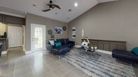 This single-floor space is defined by its striking vaulted ceilings that rise dramatically throughout the main living areas, creating a strong sense of openness and volume. The interior is finished in warm taupe and gray tones, providing a cohesive, neutral backdrop that complements the contemporary furnishings throughout.

The main lounge area is generously proportioned, accommodating multiple seating arrangements including a navy leather sectional sofa and a separate brown upholstered sofa, each anchored by abstract area rugs in blue and gray tones. A glass-top coffee table with chrome legs adds a modern touch, while curated wall art and decorative accents contribute to the polished aesthetic. Large windows and glass-paneled doors flood the space with natural light and offer views of a lush outdoor area.

A dedicated counter station with dark cabinetry, an arched architectural detail, and recessed lighting serves as a functional reception or service area, suggesting the space is suited for professional or community use. Tile flooring runs throughout, offering durability and a clean, uniform appearance. Ceiling fans with recessed lighting provide practical comfort, rounding out this well-appointed, versatile interior.