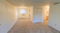 This single-floor space offers a clean and functional layout with a warm, neutral aesthetic throughout. The main living and dining area is generously sized, featuring wall-to-wall beige carpeting that adds a soft, comfortable feel underfoot. A classic chandelier hangs above the dining portion of the room, providing an elegant focal point and ample overhead lighting. Natural light filters in through a window fitted with vertical blinds, brightening the space and creating an open, airy atmosphere. A baseboard heating unit runs along the exterior wall, ensuring comfort year-round. A large closet with louvered bifold doors offers substantial built-in storage, a practical feature that enhances the overall livability of the space. The textured ceiling adds a subtle layer of architectural detail, consistent with the unit's understated, traditional character. A connecting hallway leads to additional rooms, hinting at a thoughtful floor plan that balances open communal areas with more private spaces. The overall ambiance is bright, clean, and move-in ready, making it a versatile space suited to a variety of lifestyles and furnishing styles.