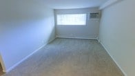 This single-floor space presents a clean, straightforward layout centered around a spacious, open living area. The room is finished with neutral-toned wall-to-wall carpeting that extends throughout, providing a soft and comfortable underfoot feel. The walls are painted in a light, neutral tone, contributing to a bright and airy atmosphere. A wide horizontal window along the far wall allows natural light to fill the room, while vertical blinds offer privacy and light control. Climate comfort is well-addressed, with a baseboard heating unit running along the exterior wall beneath the window, ensuring even heat distribution throughout the colder months. A wall-mounted air conditioning unit positioned in the upper corner of the room provides supplemental cooling during warmer seasons. The room's simple, open design offers flexibility for a variety of furniture arrangements and uses. Clean baseboard trim lines the perimeter of the room, adding a finishing touch to the otherwise minimalist interior. Overall, the space is functional and move-in ready, with practical climate control features and ample natural light.