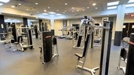 This modern fitness center offers a comprehensive workout environment designed to accommodate a wide range of exercise routines. The space is outfitted with a curated selection of Life Fitness strength training machines, including cable systems and resistance equipment, strategically arranged to allow for comfortable movement between stations. Multiple treadmills with integrated display screens line the room, providing dedicated cardio options for users of all fitness levels. The flooring features durable, dark rubberized material, ideal for high-traffic athletic use and easy maintenance. Floor-to-ceiling mirrors span the walls, visually expanding the space and allowing users to monitor their form during workouts. The ceiling is finished with a drop-tile design and features a combination of recessed lighting and skylight panels that flood the room with bright, even illumination. Ceiling fans contribute to consistent air circulation throughout the facility. The overall ambiance is clean, professional, and purposefully laid out, creating an inviting atmosphere that supports both focused individual training and general fitness activity. The open layout ensures ample space between equipment, promoting a safe and efficient workout experience.