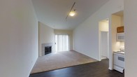 This single-floor space presents a well-proportioned living area distinguished by vaulted ceilings that enhance the sense of openness and airiness throughout. The room features a notable transition between two distinct flooring materials: dark hardwood flooring in the entry area gives way to neutral wall-to-wall carpeting in the main living space, clearly delineating functional zones within the room.

A tile-surround fireplace serves as a focal point along one wall, adding both a functional and architectural element to the space. The fireplace features classic square tile detailing and a dark firebox, contributing a traditional touch to the otherwise clean, minimalist aesthetic.

Natural light enters the space through a large sliding glass door dressed with vertical blinds, offering access to an exterior area while allowing for adjustable light control. The walls are finished in a crisp, neutral white throughout, complementing the light-toned ceiling and creating a bright, clean backdrop. Recessed track lighting and a flush-mount ceiling fixture provide overhead illumination. A doorway visible to the right suggests additional rooms beyond the main living area.