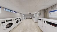 This shared laundry facility is a practical, well-organized space designed to accommodate high-volume use across two interconnected rooms. The layout is efficiently arranged, with rows of commercial-grade front-loading washing machines and top-loading dryers lining the walls on both sides, providing ample capacity for multiple users simultaneously. The flooring throughout is polished terrazzo, offering a durable and easy-to-maintain surface that contributes to the clean, functional aesthetic of the space. Ceilings feature a suspended tile grid with recessed pot lighting, ensuring consistent and even illumination throughout. The walls are finished in a neutral grey tone, giving the facility a tidy, modern appearance. One section of the space includes a built-in countertop with white cabinetry and a utility sink, providing a convenient area for hand-washing or pre-treating garments. A standalone utility sink is also present in a secondary area of the facility. A payment terminal is mounted near the entrance, and informational signage is posted on the walls. The space is accessed through a dedicated door and includes a baseboard heating unit, ensuring comfort year-round. Narrow horizontal windows along one wall allow for a degree of natural light. Overall, this laundry facility is a clean, functional, and well-maintained amenity space.