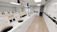 The laundry facility at Lake Castleton is a well-appointed, single-floor communal space designed with both functionality and cleanliness in mind. The room features a generous row of top-load washing machines lined along a central island, complemented by stacked dryer units positioned along the perimeter walls. Each machine is equipped with digital controls, making operation straightforward for residents.

The interior maintains a crisp, all-white color palette throughout, with the machines, cabinetry, and walls contributing to a bright, orderly atmosphere. Light wood-look tile flooring runs the length of the room, adding a warm contrast to the otherwise neutral tones. Overhead, recessed panel lighting and skylights ensure the space is evenly and thoroughly illuminated throughout the day.

The ceiling is fitted with ceiling-mounted speakers or security hardware, reflecting attention to both amenity and safety. A trash receptacle is conveniently placed near the dryer units for easy disposal. A partially visible adjoining space, glimpsed through a glass-paneled door, appears to serve as an office or business center, suggesting the laundry facility is part of a broader amenity suite within the community. Overall, the space is clean, modern, and efficiently organized.