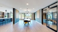 Located at 1994 Springfield Road in Kelowna, BC, this contemporary coworking space offers a thoughtfully designed, single-floor layout that balances collaborative work areas with private, focused workspaces. The open-plan main area is anchored by a sleek kitchen island with bar seating, complemented by warm wood cabinetry, open shelving, and modern dark-toned lower cabinets — a functional yet stylish hub for the space. Two distinct dining areas provide flexible seating arrangements: one features a long communal table with track lighting overhead, ideal for group work or team gatherings, while the other offers intimate booth-style seating framed by floor-to-ceiling windows that flood the area with natural light and offer views of the surrounding Kelowna streetscape. The lounge area provides a more relaxed setting for informal meetings or breaks. A dedicated meeting room with a long wooden table, upholstered chairs, and wall-mounted display offers a quieter environment for focused sessions. Private work pods with built-in desks and individual climate controls are available for heads-down work. Throughout, light hardwood flooring, neutral tones, and carefully considered lighting create a professional yet welcoming atmosphere suited to a wide range of work styles.