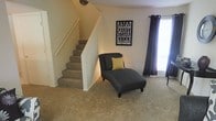 This two-story residence offers a practical and comfortable layout spread across two well-connected floors. The main living area is finished with plush wall-to-wall carpeting that adds warmth underfoot and contributes to a cozy, inviting atmosphere. A carpeted staircase with a white-painted surround and handrail provides a clean transition between the two levels, positioned conveniently within the living space.

Natural light filters into the living area through a sizable window dressed with dark curtain panels, offering both brightness and privacy control. The neutral wall tones throughout create a versatile backdrop, lending the space a calm and open feel that can accommodate a variety of interior styles.

The living area provides ample room for seating and relaxation, as evidenced by its open floor plan. Decorative wall art adds a personal touch and a sense of character to the otherwise understated aesthetic. The overall design of the home prioritizes functionality and livability, with straightforward finishes and a layout that makes efficient use of the available space on both floors. The combination of natural light, neutral tones, and soft carpeting throughout gives this residence a welcoming and move-in-ready quality.