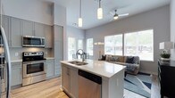 This well-designed single-floor space showcases a contemporary open-concept layout that effortlessly connects the kitchen and living areas into one cohesive, functional environment. The kitchen is a standout feature, centered around a large island with a sleek white quartz countertop, a double stainless steel sink, and a built-in dishwasher. Soft gray cabinetry lines the walls, complemented by stainless steel appliances including a range, microwave, and a subway tile backsplash that adds a clean, modern touch. Pendant lighting hangs above the island, enhancing both the aesthetics and functionality of the workspace. The soaring ceilings amplify the sense of openness throughout, while a ceiling fan in the living area ensures year-round comfort. Large windows flood the space with abundant natural light, offering views of the surrounding greenery and creating a bright, airy ambiance. The living area is generously proportioned, providing ample room for comfortable seating and relaxed everyday living. Light hardwood flooring runs throughout, tying the open-plan space together with warmth and continuity. Overall, the space strikes a refined balance between modern design and practical livability.