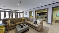 This single-floor space is thoughtfully designed to offer a comfortable and functional environment for gathering and relaxation. The main lounge area is anchored by a large, L-shaped sectional sofa in warm taupe tones, complemented by a standalone sofa and a stylish patterned ottoman that doubles as a coffee table. Accent pillows in olive green and grey add a pop of color, tying together the contemporary aesthetic. Recessed lighting illuminates the space from above, while large multi-pane windows along one wall allow generous natural light to fill the room, creating an open and inviting atmosphere. A wall-mounted flat-screen television is positioned for easy viewing from the seating area. An open archway leads directly into a well-appointed kitchen, featuring dark espresso cabinetry, a sleek countertop breakfast bar with tall bar stools, and stainless steel appliances. The kitchen's accent wall in a bold chartreuse green adds a vibrant contrast to the darker cabinetry. Light wood-tone flooring flows throughout, lending warmth and cohesion to the overall design. The layout is ideal for communal use, balancing lounge comfort with practical kitchen amenities.