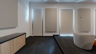 Located at 915 Division St in Nashville, TN, this thoughtfully designed single-room space serves as a premium screening or presentation lounge. The interior embraces a clean, modern aesthetic defined by crisp white walls, dark charcoal carpet flooring, and a minimalist sensibility throughout.

The room is anchored by a large wall-mounted display screen, ideal for presentations, screenings, or collaborative viewings. Seating is arranged in a theater-inspired layout, combining plush cream-toned curved accent chairs and a generous tufted sectional sofa with cognac leather ottoman accents — creating a comfortable yet sophisticated atmosphere.

Acoustic treatment is a defining architectural feature, with large fabric sound panels mounted both on the walls and ceiling, ensuring optimal audio performance. Recessed ceiling lighting provides even, adjustable illumination throughout the space. A subwoofer is positioned near the screen wall, underscoring the room's audio-visual capabilities.

Near the entry, floor-to-ceiling windows fitted with roller shades allow natural light to be precisely controlled, while illuminated door frames add a refined, modern touch. A built-in credenza provides functional storage near the entrance. The space is accessible via a set of sleek black double doors, completing its polished, professional character.