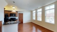 This two-floor space combines practical design with warm finishes throughout. The kitchen is a central highlight, featuring rich medium-toned wood cabinetry that extends upward toward a vaulted ceiling, maximizing storage. Dark granite-style countertops provide a striking contrast, while a double stainless steel sink sits beneath a classic pendant light. Black appliances, including a refrigerator and range, add a cohesive, polished aesthetic. A louvered bi-fold door offers convenient pantry or utility storage, and a peninsula counter creates a natural division between the kitchen and the main living area. The living space is bright and open, benefiting from large, multi-pane windows that invite abundant natural light and views of the surrounding streetscape. Rich hardwood flooring flows throughout, adding warmth and continuity to the space. Neutral-toned walls keep the interior feeling clean and versatile. The overall ambiance is inviting, blending functional kitchen design with a spacious, light-filled living area that is well-suited for everyday living across two floors.