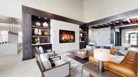 Located in Plainfield, Illinois, this single-floor community clubhouse at 13915 S Autumn Way blends contemporary design with welcoming, functional spaces. The centerpiece of the facility is a sophisticated lounge area boasting soaring ceilings, a striking white stacked-stone fireplace flanked by dark wood built-in shelving with integrated lighting, and a curated mix of leather and upholstered seating arranged around a rich wood coffee table. The open layout connects seamlessly to additional seating areas, creating an inviting social atmosphere. The fully equipped kitchen features light wood cabinetry reaching to the ceiling, stainless steel appliances including a side-by-side refrigerator and wall oven, a large white quartz island with bar seating, subway tile backsplash, and industrial-style pendant lighting. An abundance of natural light pours in through oversized windows. The dining area accommodates a large gathering table and chairs beneath a handsome dark wood ceiling detail, with floor-to-ceiling sliding glass doors opening directly to an outdoor pool and patio area. Throughout the space, wide-format tile flooring, dark structural columns, and thoughtful architectural detailing create a cohesive, upscale aesthetic.