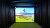 This purpose-built indoor golf simulator room offers a well-appointed and immersive experience for golf enthusiasts. The space is anchored by a High Definition Golf simulator system, complete with a large, high-resolution projection screen framed by floor-to-ceiling black drape curtains that enclose the hitting bay and minimize light interference. The floor transitions from light hardwood-style flooring in the lounge area to lush artificial turf on the hitting surface, which features a distinct putting green insert for added realism. Ceiling-mounted projectors and precision tracking cameras are strategically positioned overhead to deliver accurate shot simulation. A wall-mounted monitor and dedicated control station — housing the simulator's computer hardware and keyboard — allow for easy course selection and system management. The ceiling incorporates a circular ventilation unit and recessed lighting, maintaining a clean, modern aesthetic throughout. On the opposite end of the room, a relaxed seating area with plaid accent chairs and a round side table provides a comfortable space for guests to observe and unwind. Framed artwork adorns the white walls, adding a polished finishing touch. The overall ambiance is sleek, functional, and purpose-driven, making it an ideal private facility for practice, entertainment, or competitive play.