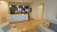 This single-floor residence offers a thoughtfully designed open-concept layout that seamlessly connects the kitchen and living areas. The kitchen is a standout feature, outfitted with dark espresso cabinetry, stainless steel appliances including a French door refrigerator with ice dispenser, a built-in microwave, and a gas range. A subway tile backsplash adds a clean, modern touch, while light granite countertops provide generous prep space. The kitchen island serves as both a functional workspace and a casual dining area, complemented by sleek acrylic bar stools. Pendant lighting hangs above the island, while a track lighting system illuminates the cooking space. The flooring throughout the main area features polished concrete, lending an industrial-contemporary feel that is both durable and visually striking. A natural fiber area rug in the living area adds warmth and texture, grounding the comfortable seating arrangement. The space benefits from an open, airy flow, with interior doors leading to additional rooms beyond the main living area. A chalkboard accent wall near the hallway adds a creative, personalized touch. Overall, the space balances modern aesthetics with everyday functionality.