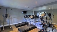 This fitness center is a purpose-built exercise space designed to support a well-rounded cardio workout routine. The room is finished with neutral gray walls and smooth hardwood-style flooring, creating a clean and motivating environment. A row of modern treadmills lines the center of the space, complemented by an elliptical trainer, providing a range of cardio options for users of varying fitness levels. A large wall-mounted mirror spans one side of the room, visually expanding the space and allowing users to monitor their form during workouts. Recessed lighting in the ceiling ensures the area is well-lit, while a wall-mounted flat-screen television adds an element of entertainment to keep workouts engaging. A window allows natural light to filter into the space, contributing to a bright and energizing atmosphere. The layout is thoughtfully organized to maximize floor space and ease of movement between equipment. Overall, this fitness center offers a practical and comfortable setting for daily exercise.