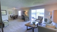This single-floor residence features an open-concept layout that seamlessly integrates the living and dining areas into one cohesive, light-filled space. The living area is carpeted throughout and anchored by a rustic wood coffee table, complemented by a mix of patterned and solid upholstered seating that adds visual interest. Large sliding glass doors dominate one wall, flooding the interior with natural light and providing direct access to a private balcony overlooking a serene waterway framed by lush tree coverage — a standout feature of the space.

The dining area is positioned near a window on the adjacent wall, offering additional natural light and a relaxed setting for meals. A transitional console table with a decorative lamp serves as a subtle divider between the living and dining zones, adding warmth and definition without interrupting the open flow.

The textured ceiling adds a layer of architectural character, while the neutral wall tones and carpeted floors contribute to a calm, cohesive ambiance throughout. The combination of water views, abundant natural light, and an open, functional layout makes this a comfortable and inviting living environment.